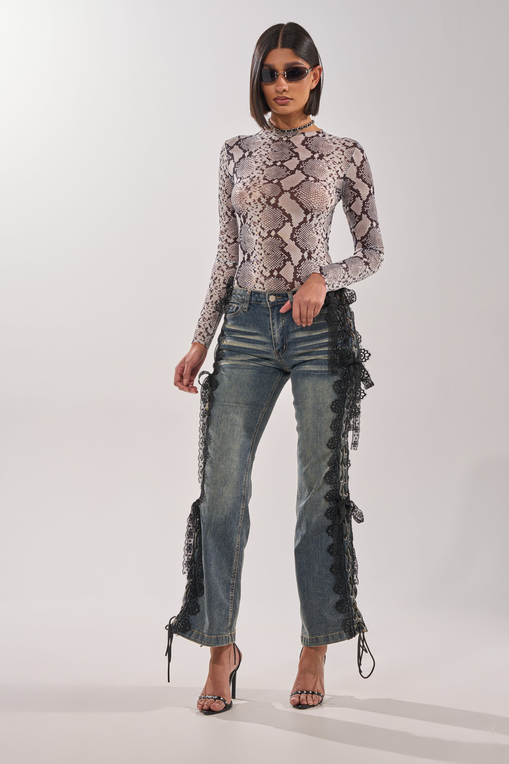 A woman poses against a plain background wearing sunglasses, strappy heels, blue jeans with black lace sides, and the OMBRE SNAKE LONG SLEEVE MESH TOP IN GREY MULTI. 7