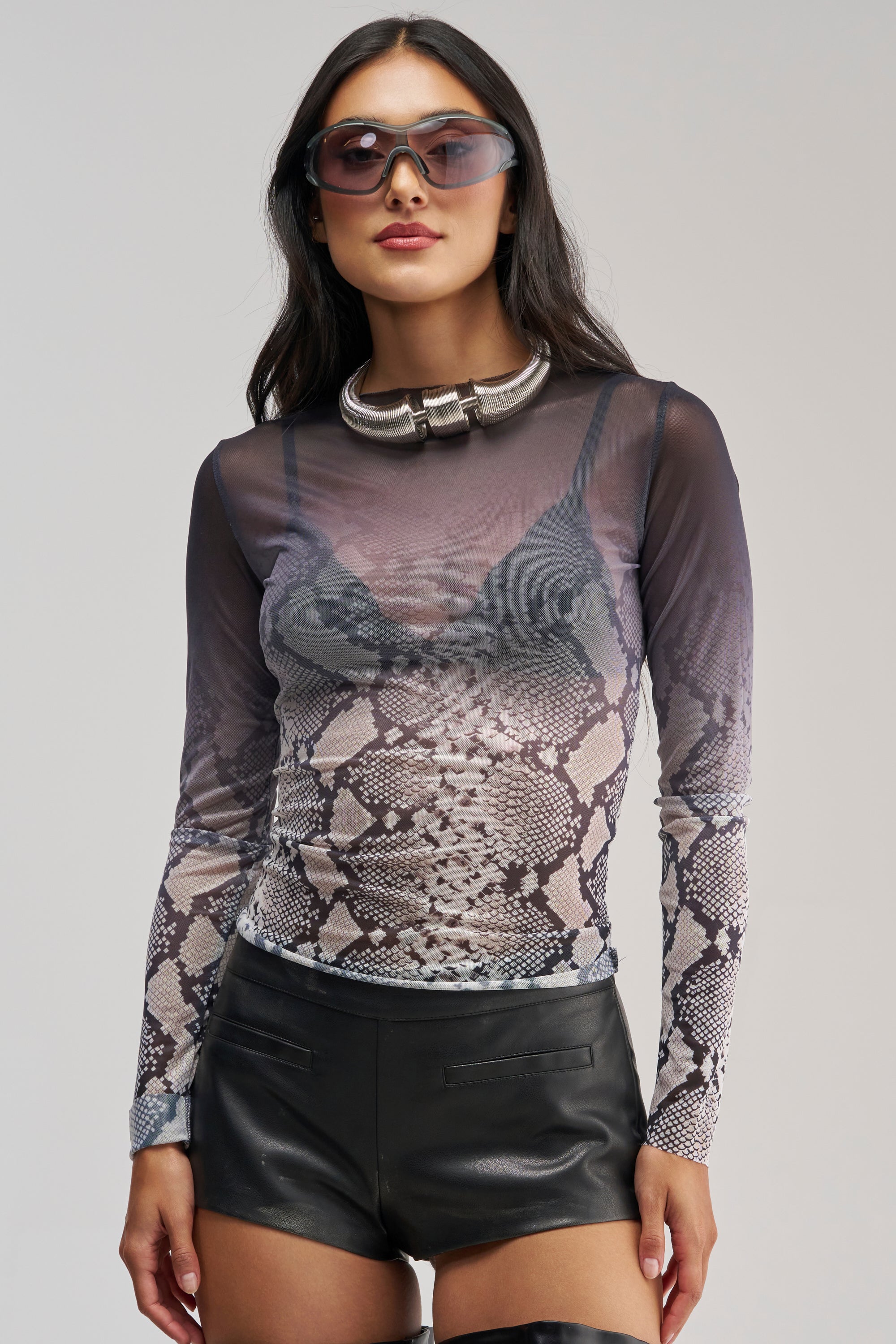 A woman wearing sunglasses, a chunky necklace, the SNAKE EYES OMBRE MESH LONG SLEEVE TOP over a black bra, and black leather shorts poses confidently against a plain background.