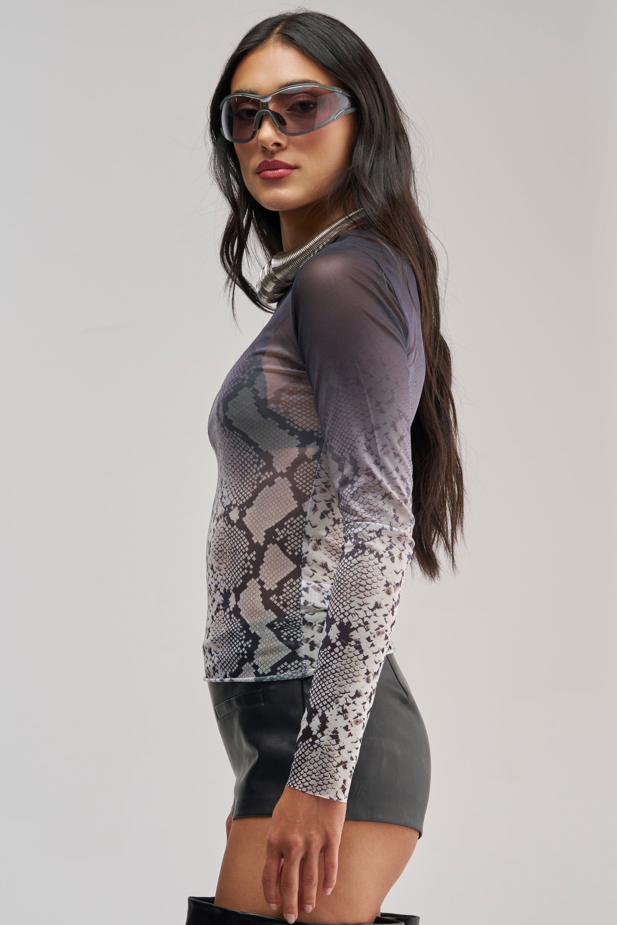 A woman wearing sunglasses, black shorts, and black knee-high boots stands in profile against a light background, showcasing the SNAKE EYES OMBRE MESH LONG SLEEVE TOP.