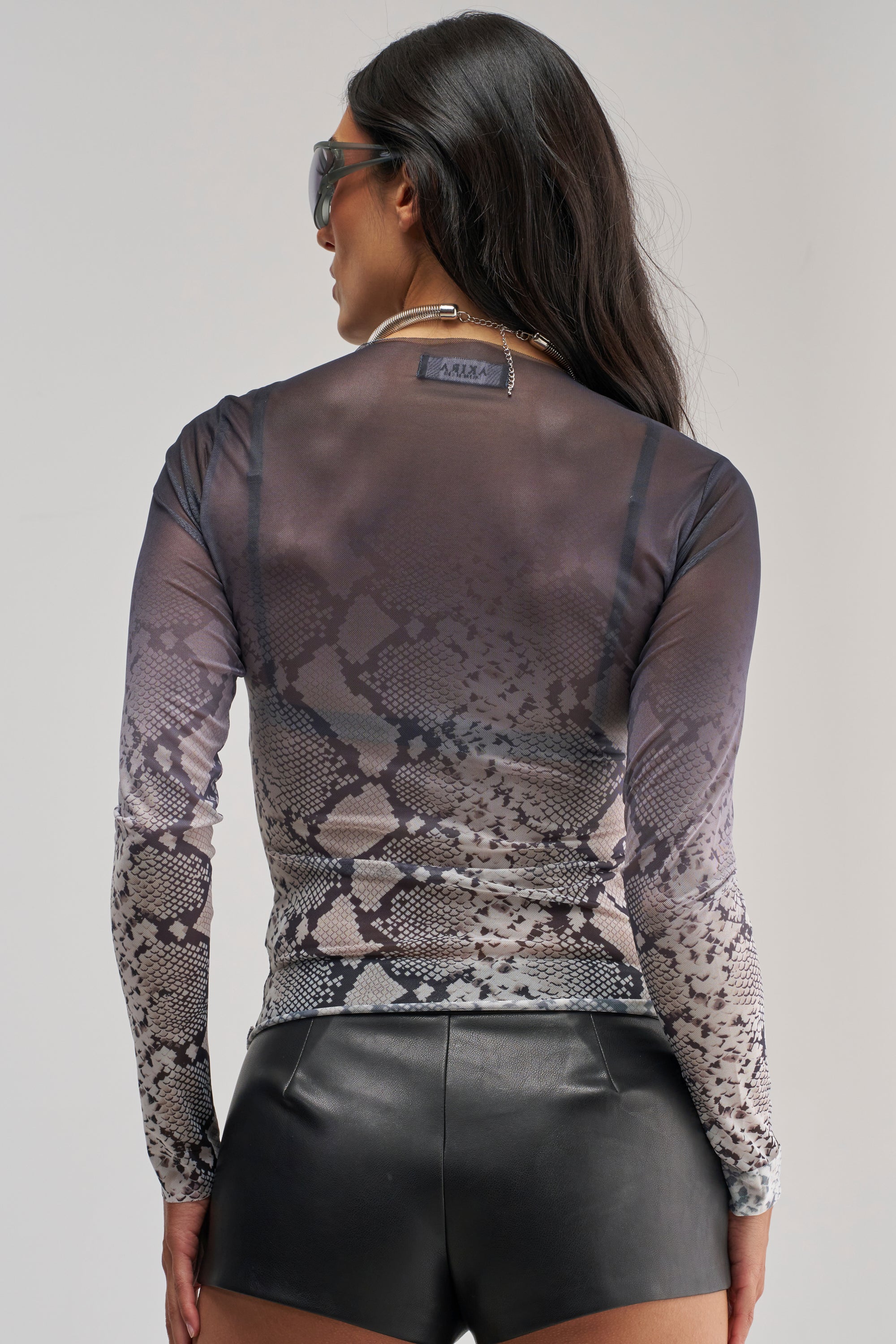 A woman wearing sunglasses, the SNAKE EYES OMBRE MESH LONG SLEEVE TOP, and black leather shorts stands facing away from the camera against a plain background.