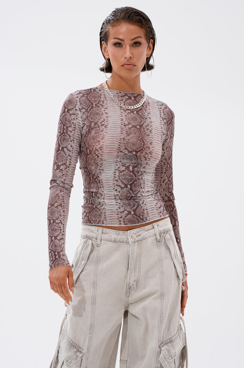 A woman with slicked-back hair wears the SNAKE CHARMER LONG SLEEVE MESH TOP and light cargo pants, accessorized with a chunky silver chain necklace, standing against a plain white background.