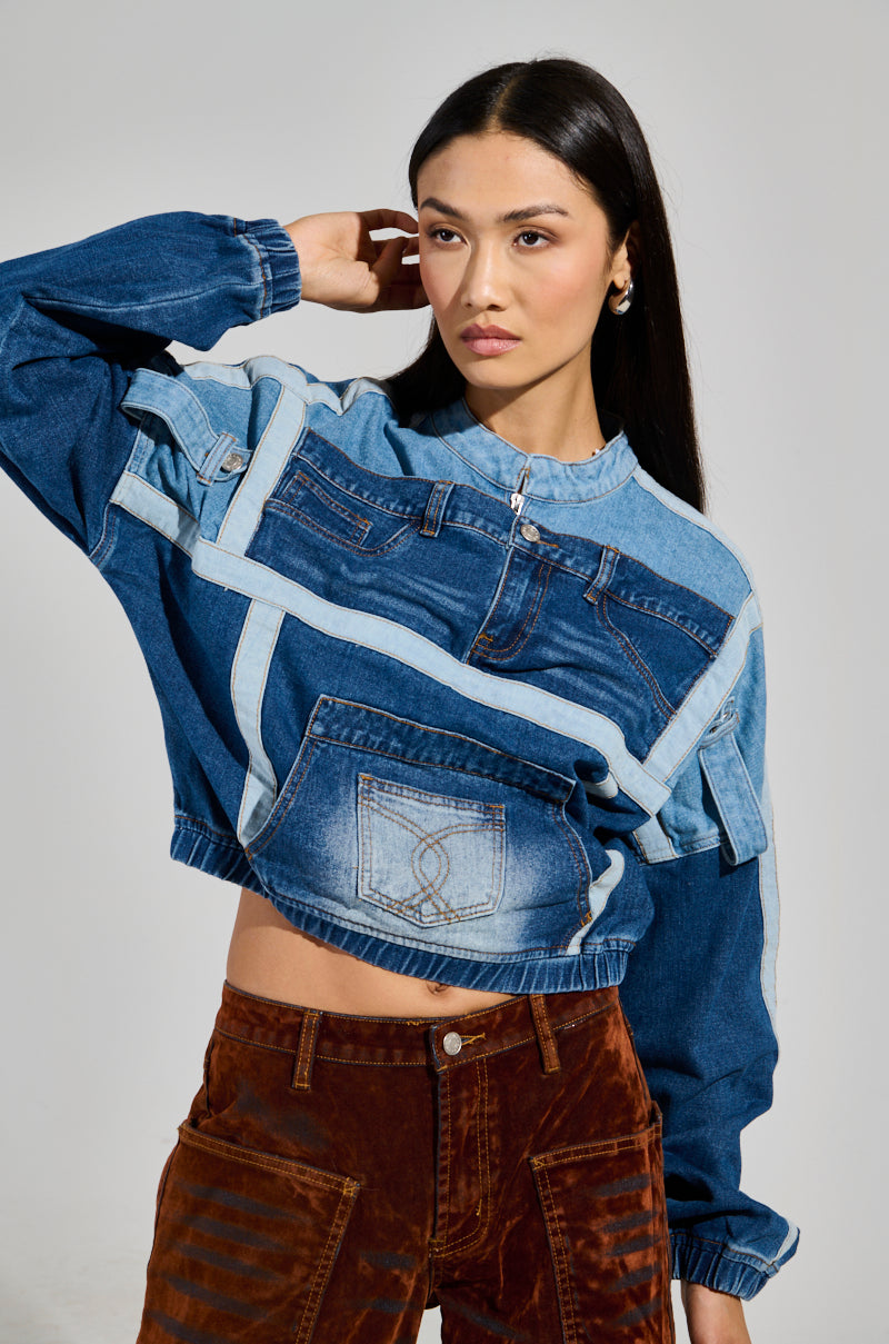 A person with long dark hair is modeling the AUBREY DENIM PULL OVER SWEATSHIRT, featuring wide sleeves and a front pocket. It is paired with brown pants to accentuate the color block design, as they pose gracefully with one hand touching their hair against a plain gray background.