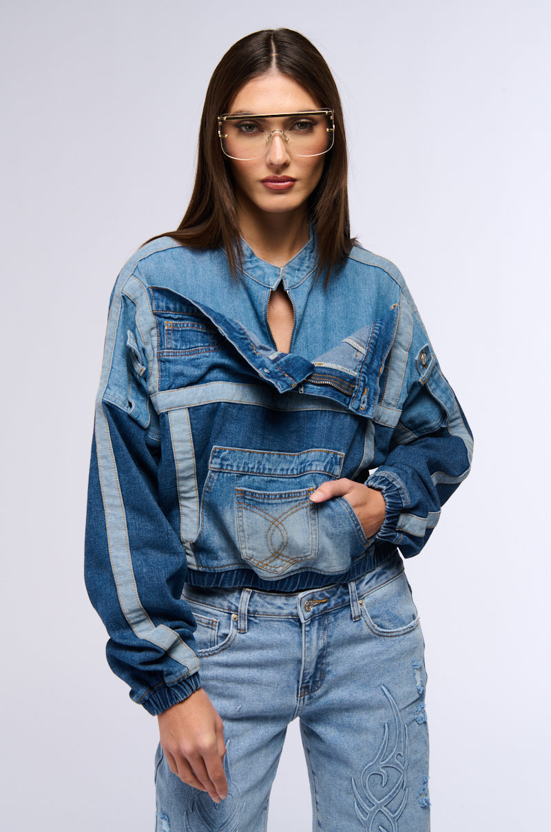 A person with long hair is wearing large glasses and a trendy AUBREY DENIM PULL OVER SWEATSHIRT, styled with multiple pockets. They seamlessly coordinate it with color block construction denim pants that feature embroidered details, standing confidently against a plain background.