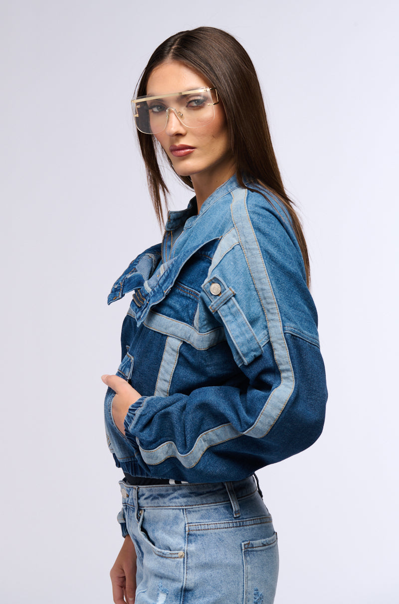 A person wearing an Aubrey denim pull-over sweatshirt and jeans stands against a plain background. They sport clear protective glasses, have straight dark brown hair, and their expression is neutral.