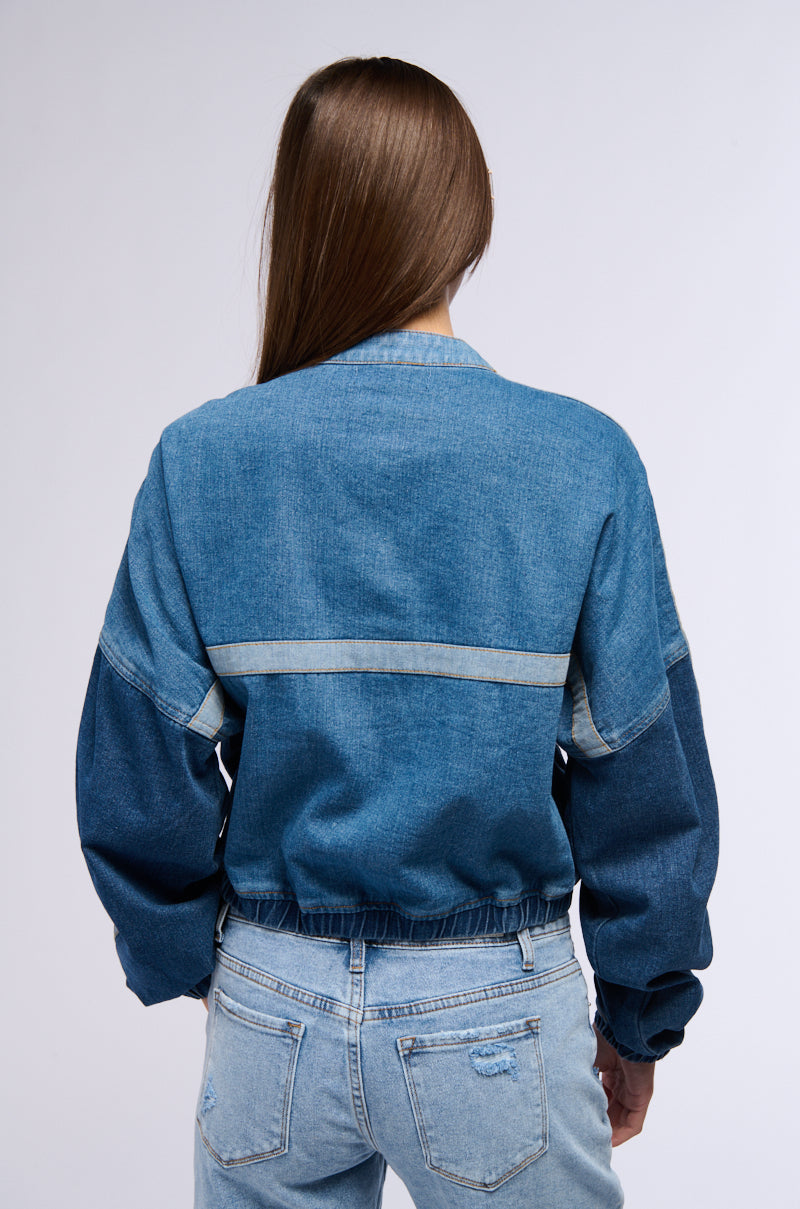 Back view of a person with long brown hair wearing the AUBREY DENIM PULL OVER SWEATSHIRT paired with light blue jeans. The sweatshirt, resembling a color block construction, features a darker blue stripe across the upper back and has an elastic waistband. The background is plain and light gray.