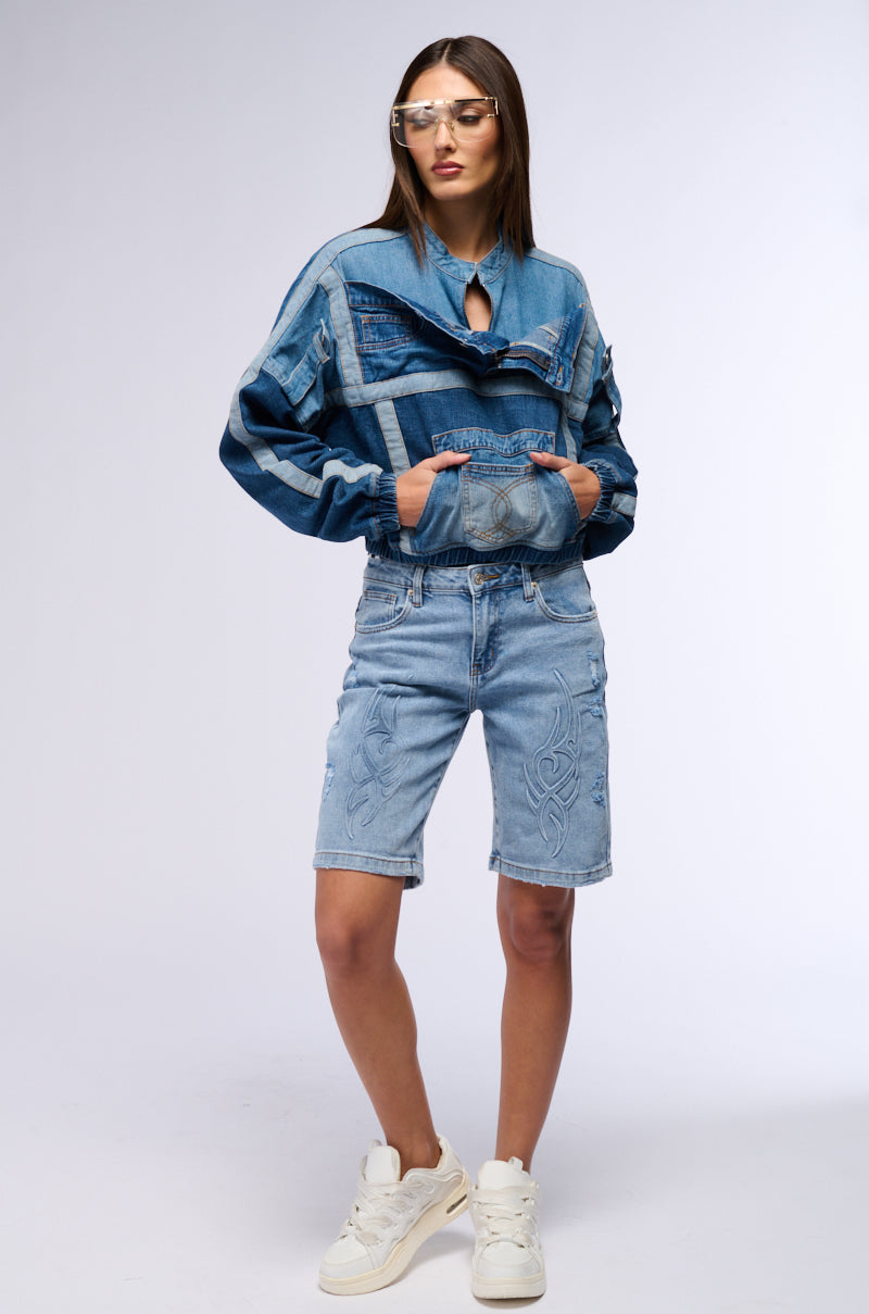 A woman poses against a plain background wearing a fashionable denim ensemble. She rocks a color block construction with her AUBREY DENIM PULL OVER SWEATSHIRT and light blue knee-length shorts, complemented by white sneakers and futuristic sunglasses. Her hands are tucked casually in her sweatshirt pockets.