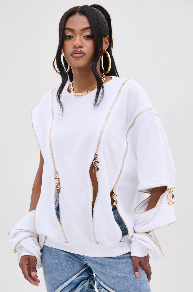 A fashion-forward woman with long black hair confidently wears an AKIRA Label SABRINA ALL OVER ZIPPER SWEATSHIRT with gold zippers and large holes, styled with blue jeans. She completes her look with large gold hoop earrings, a gold necklace, and a confident expression.