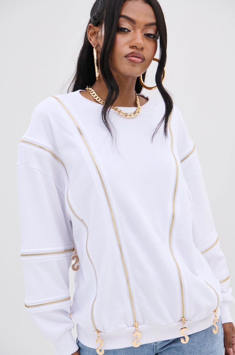 A person with long hair wears a SABRINA ALL OVER ZIPPER SWEATSHIRT in white, adorned with gold accents and pendants. They pair it with hoop earrings and a chain necklace, giving off a fashion-forward vibe and wearing a confident expression against a plain light gray background.