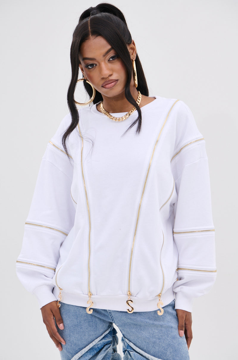The fashion-forward woman wears the AKIRA Label SABRINA ALL OVER ZIPPER SWEATSHIRT, an oversized white sweatshirt featuring gold zippers and a chain design. She stylishly pairs it with light blue jeans, a high ponytail, large hoop earrings, and a chunky gold necklace against a plain background.