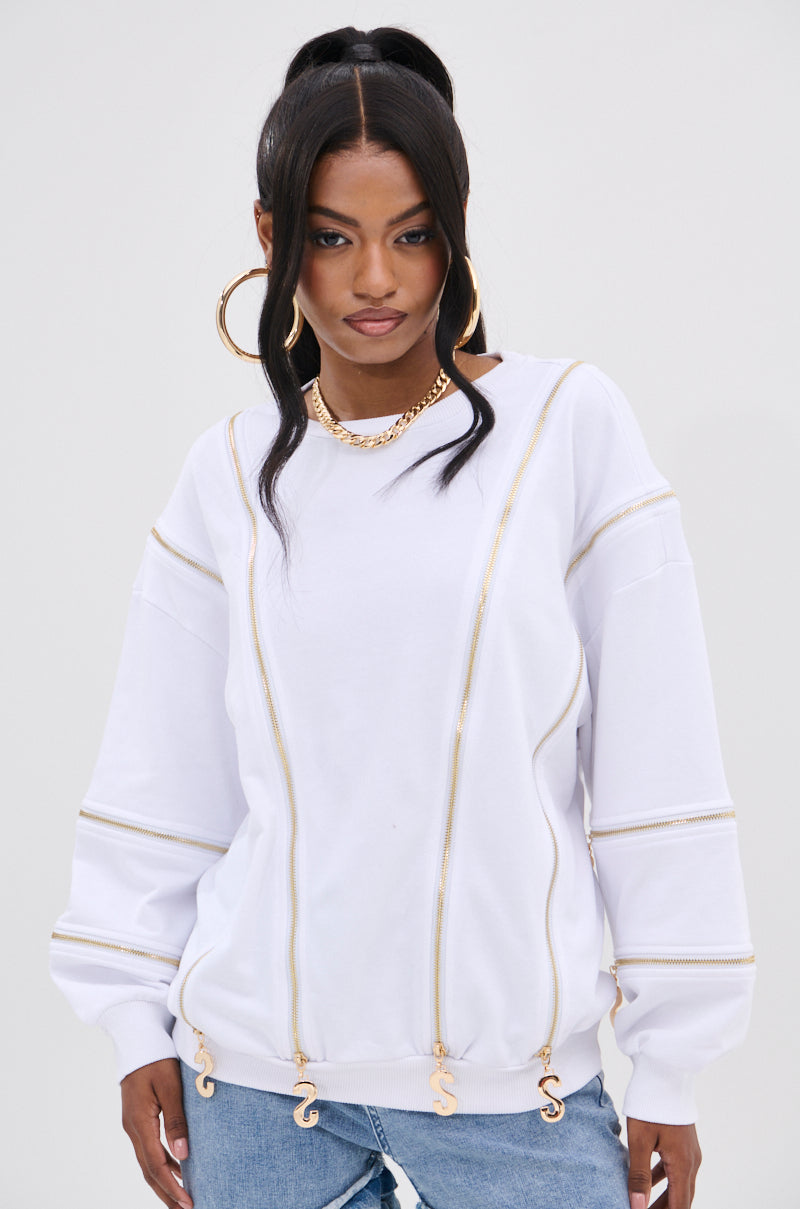 A fashion-forward individual wears the AKIRA Label's SABRINA ALL OVER ZIPPER SWEATSHIRT in white, featuring gold zippers and dollar sign pendants. With long dark hair tied up and accessorized with gold hoop earrings, a necklace, and light blue jeans on a plain backdrop, the ensemble exudes style.