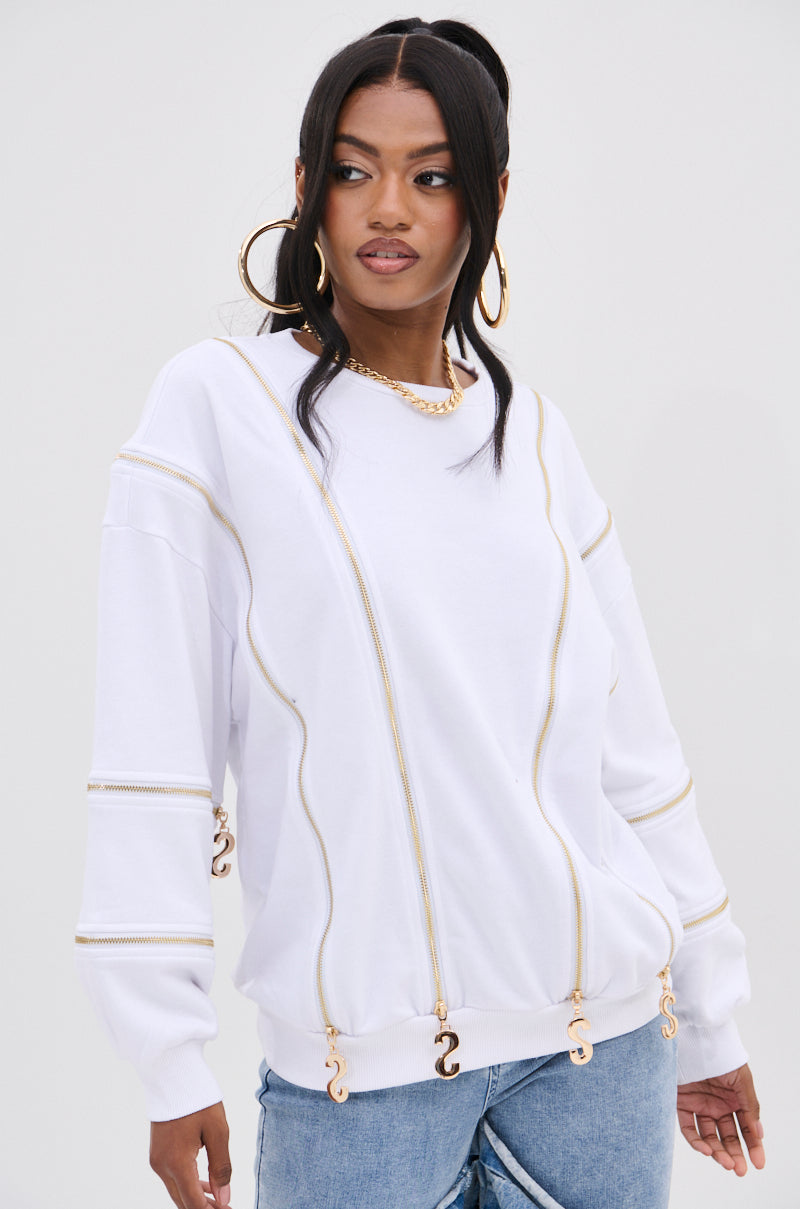 A fashion-forward individual poses against a plain backdrop, wearing the SABRINA ALL OVER ZIPPER SWEATSHIRT with gold zippers and embellishments. Their long dark hair is styled in two ponytails, accessorized with large hoop earrings, a gold necklace, and light blue jeans.