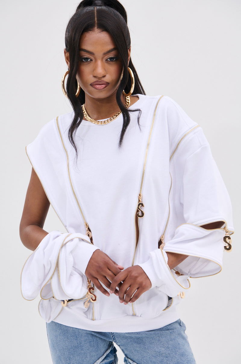 A fashion-forward woman wears the stylish AKIRA Label SABRINA ALL OVER ZIPPER SWEATSHIRT with gold zippers, paired with large hoop earrings. Her dark hair in a high ponytail complements her light blue jeans against the plain background.