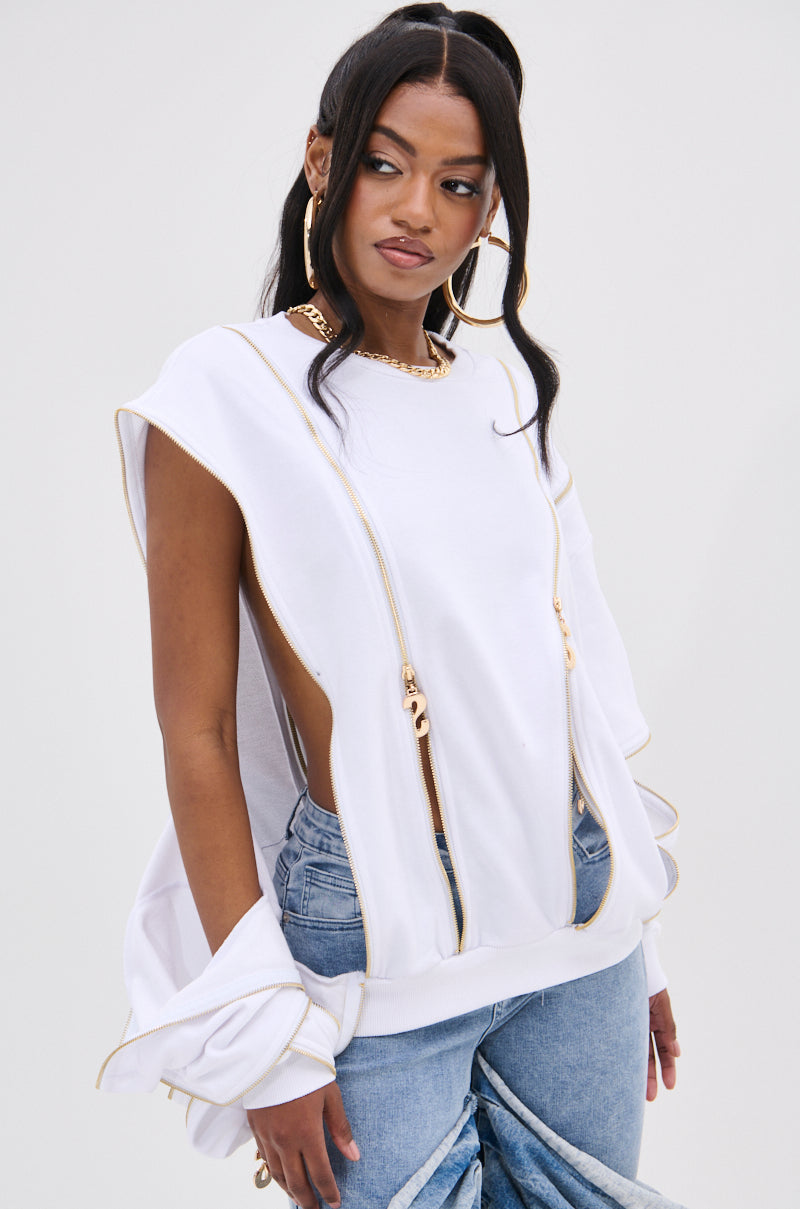 Dressed fashionably in the AKIRA Label SABRINA ALL OVER ZIPPER SWEATSHIRT, she pairs a white top featuring gold zippers with blue jeans. Her long, dark hair flows gracefully with two strands in front, complemented by large hoop earrings and a gold necklace.