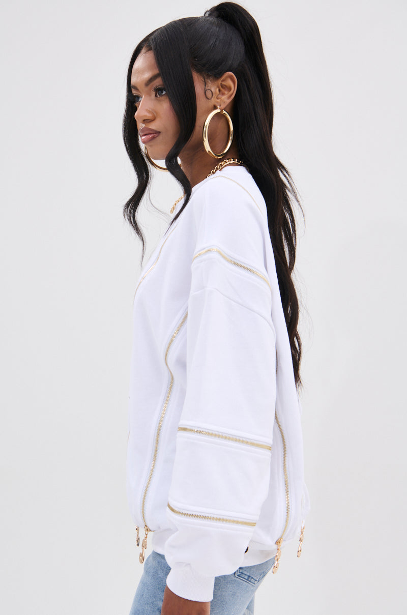 A stylish person with a long black ponytail wears large gold hoop earrings, the AKIRA Label's SABRINA ALL OVER ZIPPER SWEATSHIRT, and light blue jeans. They look to the left against a light gray background.
