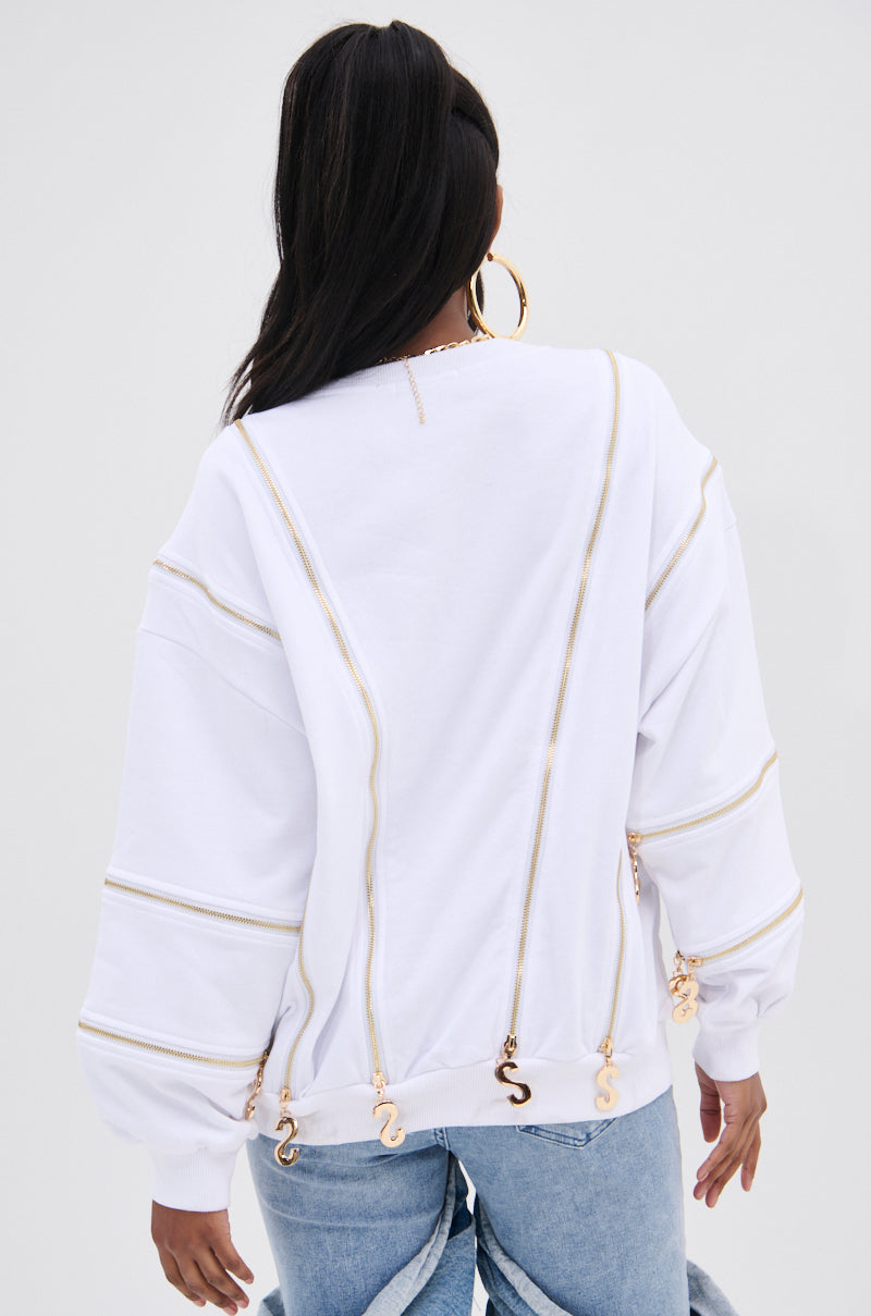 A fashion-forward individual with long dark hair, facing away, wears the AKIRA Label's SABRINA ALL OVER ZIPPER SWEATSHIRT in white with gold accents and charms. It's paired with light blue jeans and large hoop earrings against a light gray background.