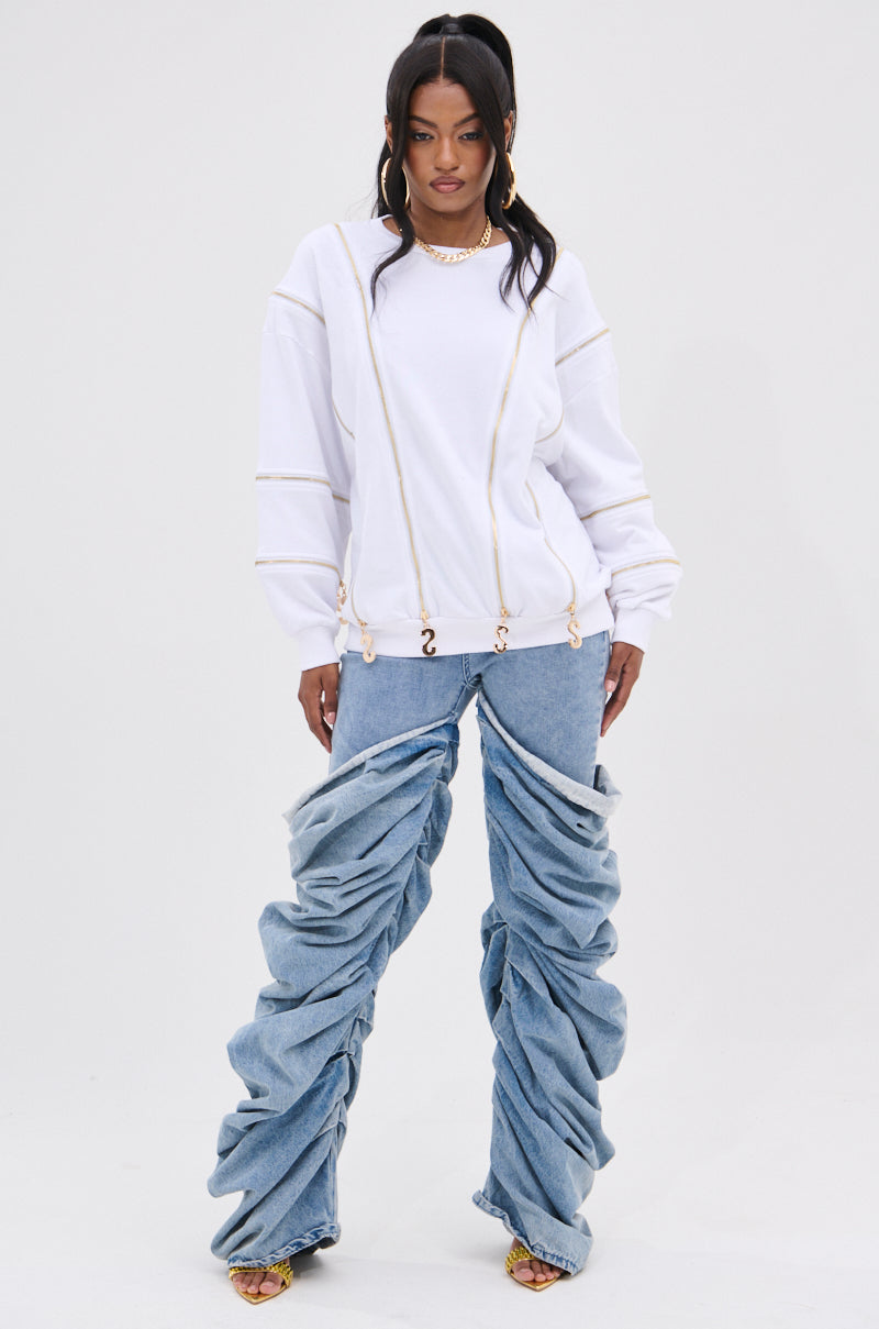 A person with long dark hair stands confidently against a plain background, showcasing a fashion-forward look. They wear a white SABRINA ALL OVER ZIPPER SWEATSHIRT paired with unique blue jeans featuring exaggerated ruched legs. Open-toed shoes and a chunky necklace complete the ensemble.
