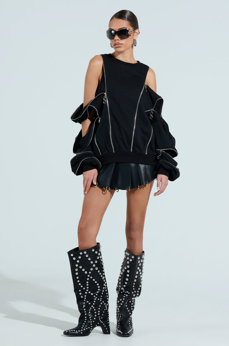 A woman in an oversized silhouette wears a chic black ensemble. Her sleeveless top, paired with the UNZIP ME SWEATSHIRT featuring silver metallic zipper details, complements the pleated skirt. Knee-high boots, sunglasses, and earrings complete her look against a plain white background.