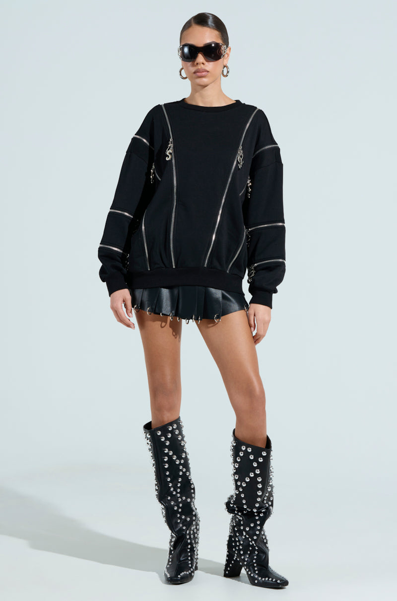 Someone wearing an UNZIP ME SWEATSHIRT with silver zippers is paired with a black leather mini skirt and knee-high studded boots. Large sunglasses and hoop earrings complete the chic look against a plain background.
