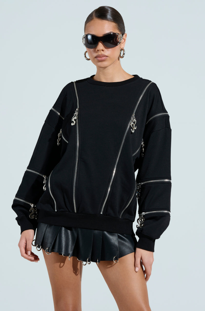 Wearing an UNZIP ME SWEATSHIRT with metallic zippers, black pleated skirt, and large sunglasses, the individual strikes a stylish pose against a plain background. Hoop earrings and sleek hair add to the chic look.