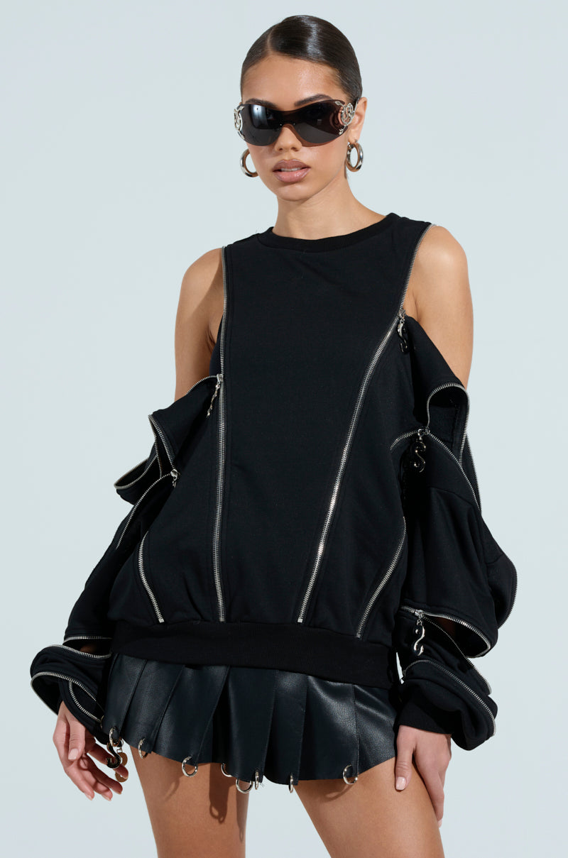 A sleek-haired person wears an outfit with the UNZIP ME SWEATSHIRT, featuring silver metallic zipper detailing, a black leather pleated skirt, oversized sleeves, large sunglasses, and hoop earrings against a light background.