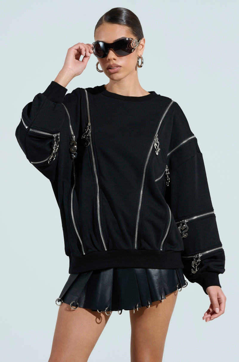 Wearing a black UNZIP ME SWEATSHIRT with silver zipper details and an oversized fit, paired with a black leather skirt. Sporting short, slicked-back hair, large sunglasses, and hoop earrings against a plain light gray background.