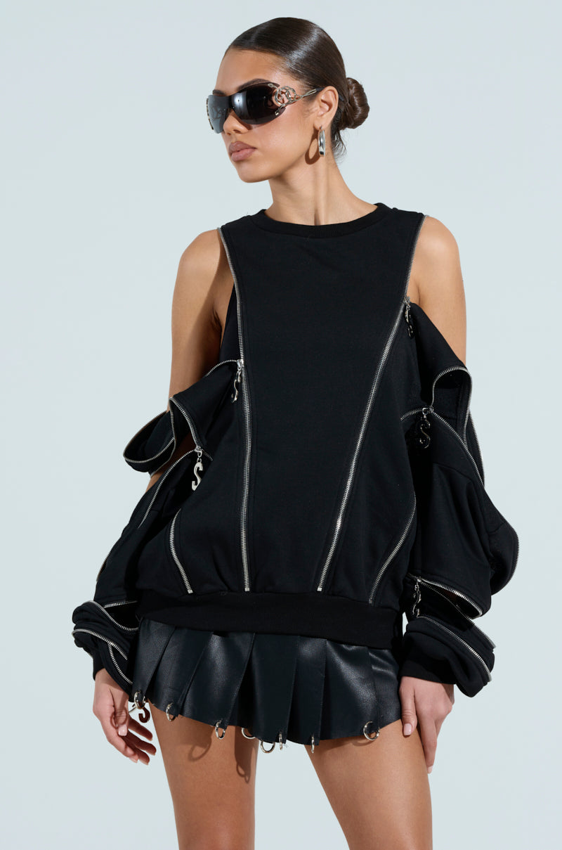 A person in a UNZIP ME SWEATSHIRT, featuring black cold-shoulder design and silver metallic zipper details, paired with a black pleated skirt. They accessorize with sunglasses and earrings, set against a light blue background.
