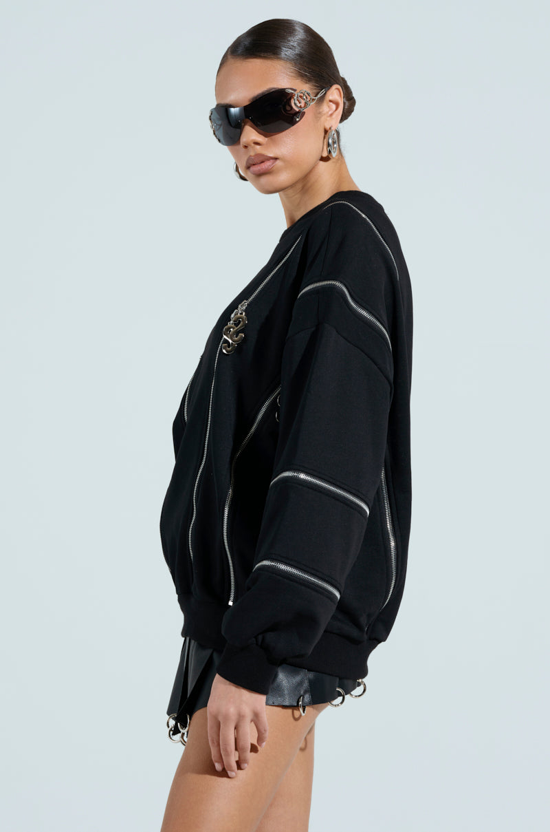 A long-haired individual in large sunglasses and hoop earrings confidently presents an oversized look with the black UNZIP ME SWEATSHIRT, showcasing silver metallic zipper details, paired with chic black shorts against a solid light backdrop.