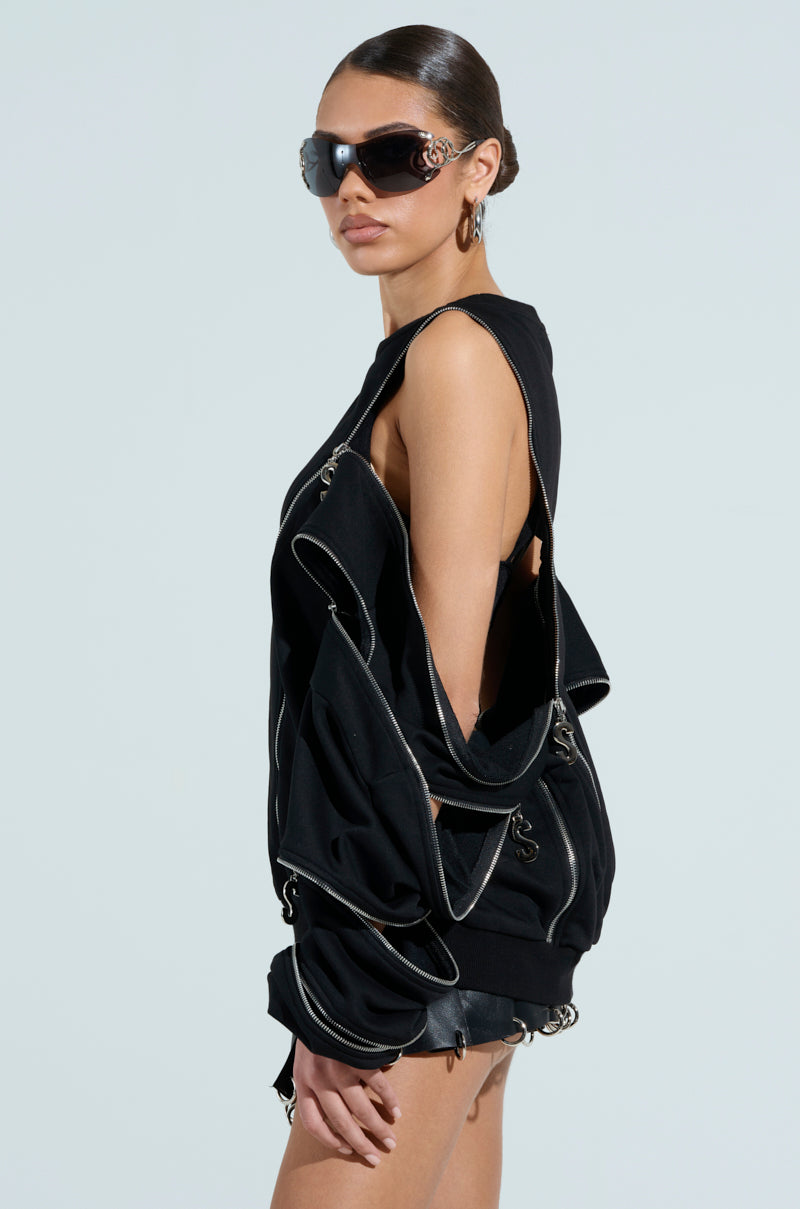 A person showcases oversized, trendy black sunglasses paired with the UNZIP ME SWEATSHIRT—a distinctive black piece featuring silver metallic zipper details. With their hair sleekly styled back, they stand against a plain light gray background.