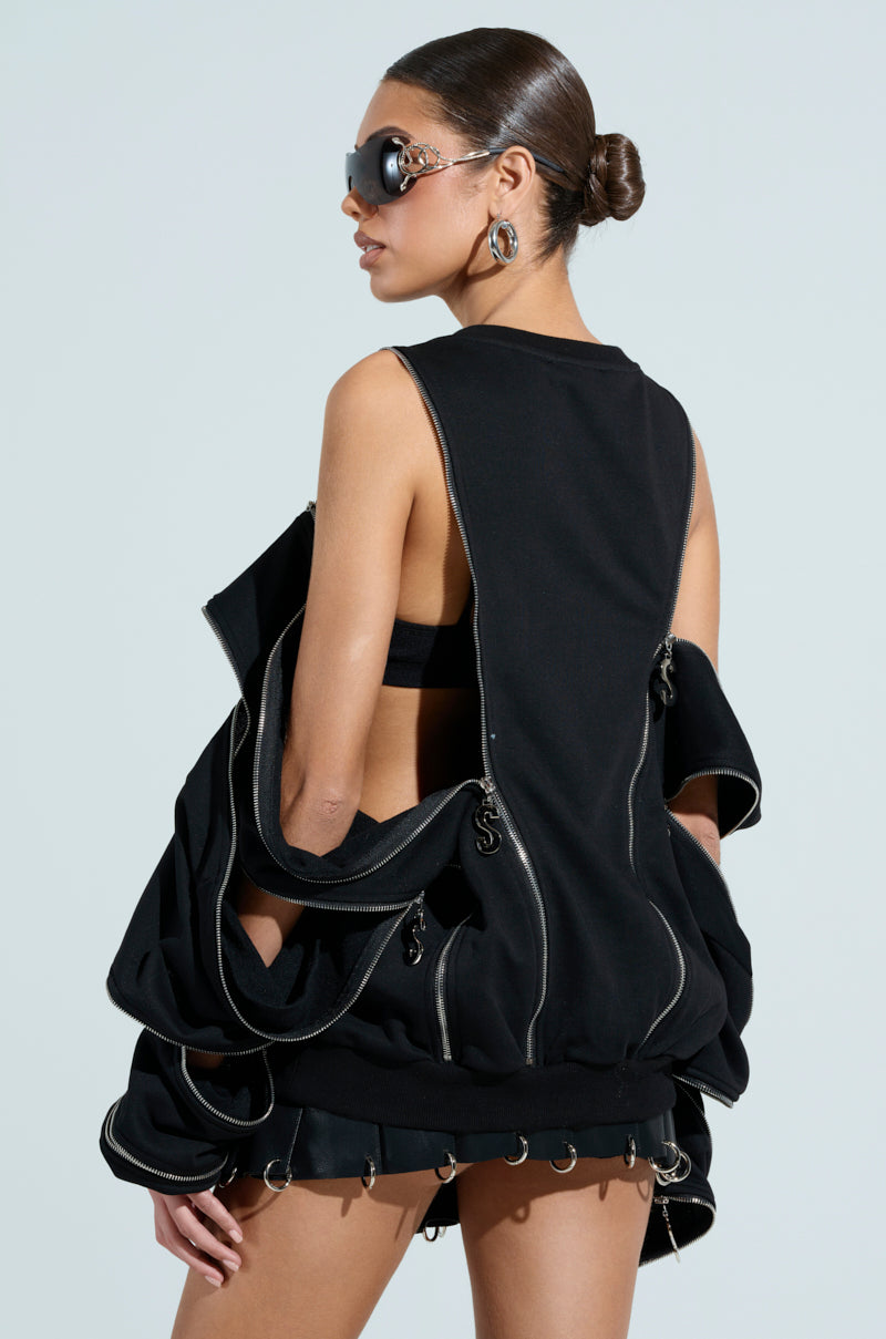 A dark-haired woman in a bun and sunglasses wears an avant-garde black UNZIP ME SWEATSHIRT with an oversized silhouette, silver metallic zipper detailing on a sleeveless top, and a skirt with metal rings, effortlessly highlighting the ensemble's back and side.
