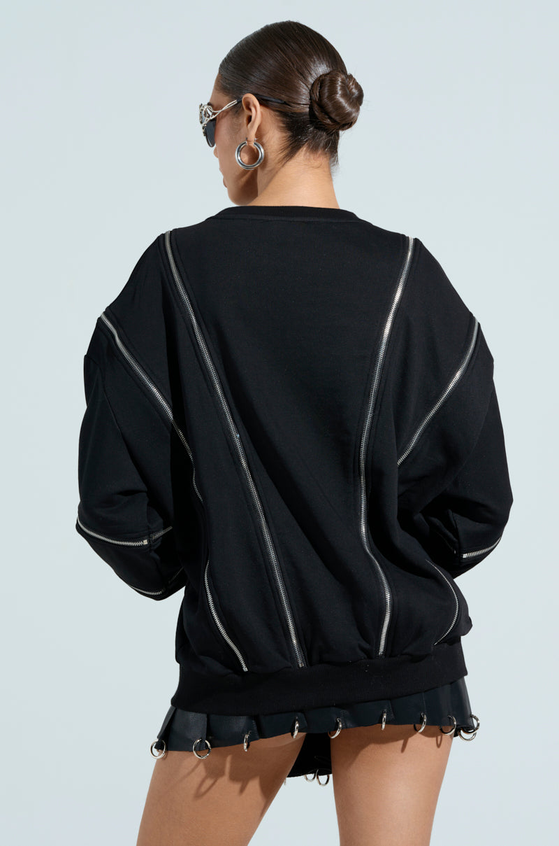 Against a light blue background, a person with a bun sports the UNZIP ME SWEATSHIRT in black, featuring silver zippers. They pair it with black shorts accented by metal rings.