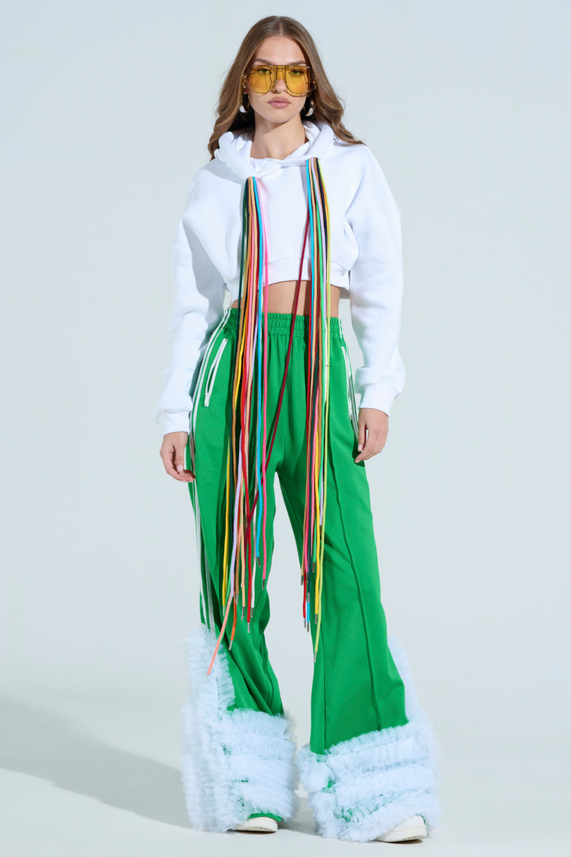 A person confidently wears the STRINGED HOODIE, a white cropped sweatshirt with adjustable drawstrings and colorful ribbons. They pair it with green wide-leg pants featuring fluffy white cuffs and yellow-tinted sunglasses, hands in pockets and long brown hair flowing down.