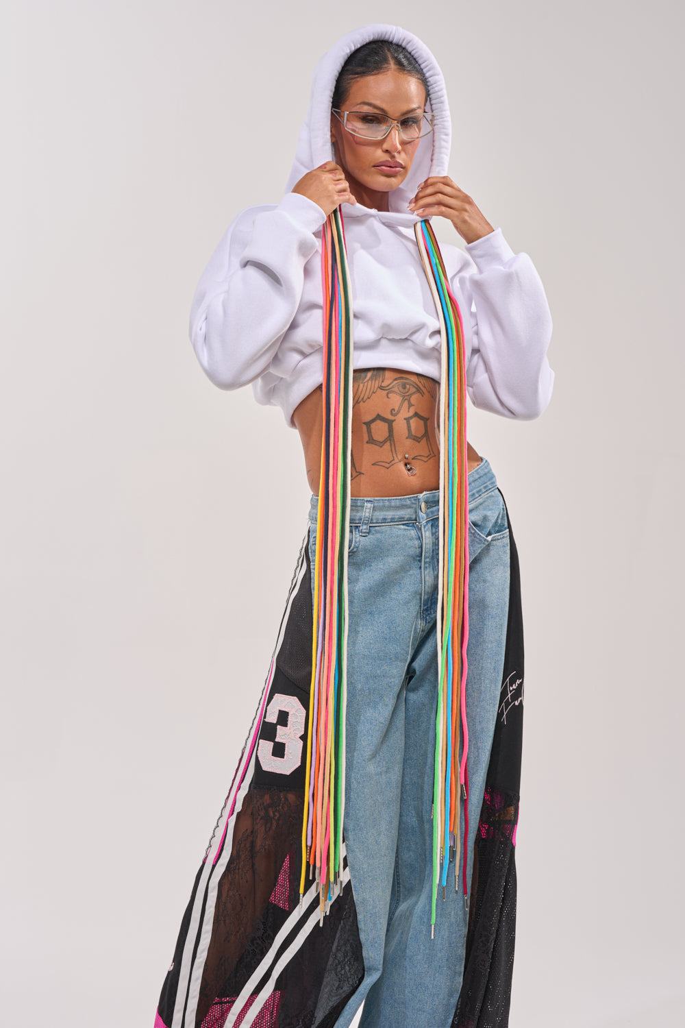 Someone stands confidently against a plain light background, wearing the STRINGED HOODIE—a cropped rainbow hoodie with a vibrant drawstring—paired with blue jeans featuring mesh and graphic details, clear glasses, and visible tattoos. 0