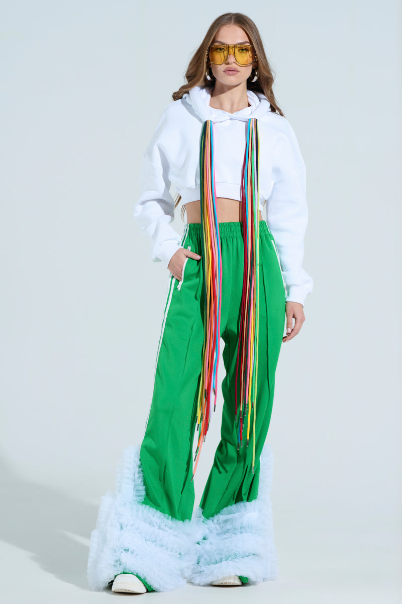 A model showcases the STRINGED HOODIE, a white cropped sweatshirt with long multicolored ribbon accessories and adjustable drawstrings. She's paired it with green wide-leg pants featuring white stripes and fluffy blue hem accents, completed by yellow-tinted sunglasses for a bold look.