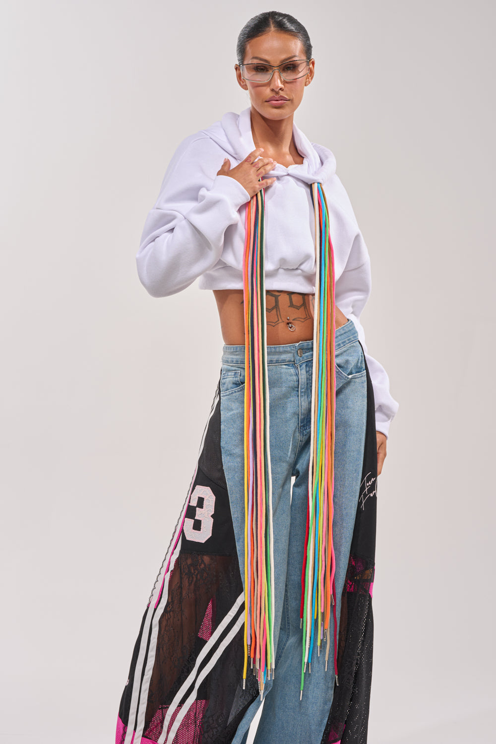 Wearing glasses and the STRINGED HOODIE with white lace details, blue jeans featuring black and pink designs, a long multicolored fringe scarf, and showing tattoos on their torso, a person poses confidently. 1