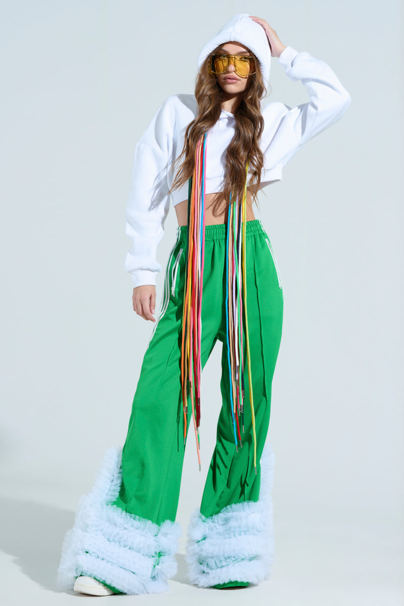 Wearing the STRINGED HOODIE, a cropped white hoodie with colorful ribbons and adjustable drawstrings, paired with green pants and white fluffy leg warmers, a person confidently poses against a plain background. Yellow-tinted sunglasses add to their bold look.