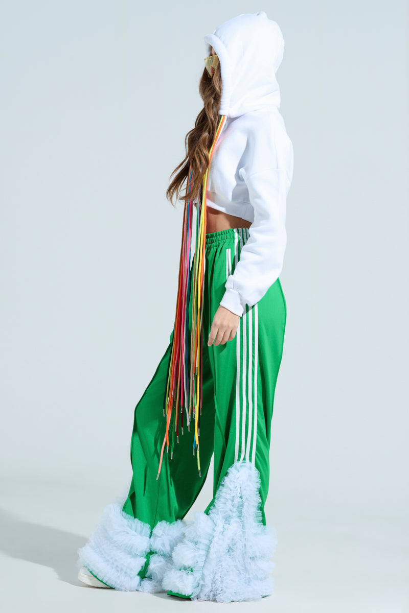 A person in a STRINGED HOODIE with colorful ribbon face covering, green track pants featuring white stripes, and furry white embellishments on the pants stands against a light background.