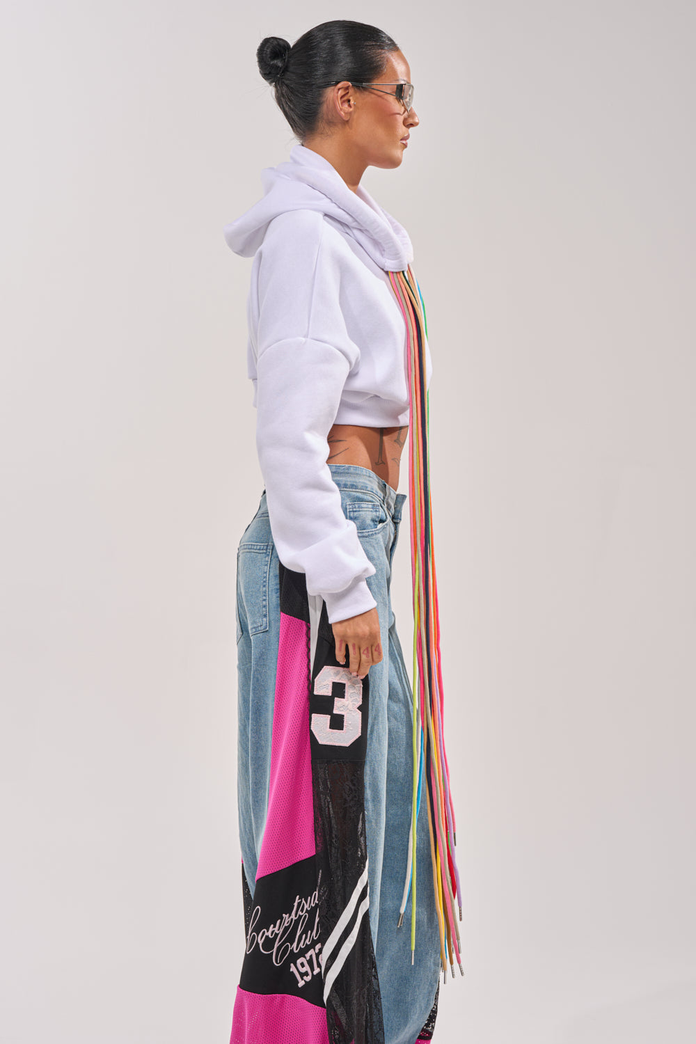 A woman in profile wears the STRINGED HOODIE, a cropped rainbow style with long multicolored strings, paired with blue wide-leg patchwork jeans. She has her hair in a bun, dons sunglasses, and poses against a plain background. 3