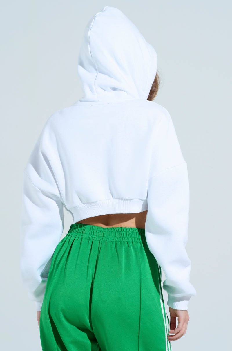 A person in a STRINGED HOODIE, a white cropped sweatshirt with an adjustable drawstring hood, and green pants with white side stripes stands back to the camera against a light blue background, creating a stylish ensemble.