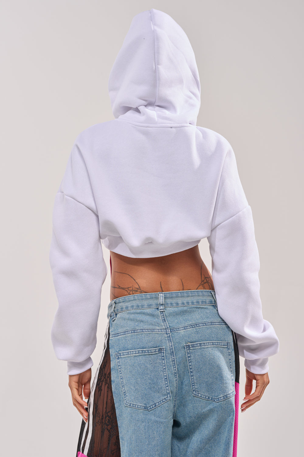 A person shown from the back wears the STRINGED HOODIE—a cropped rainbow hoodie with the hood up—and light blue jeans. Tattoos are visible on their lower back above the jeans, against a plain, light background. 4