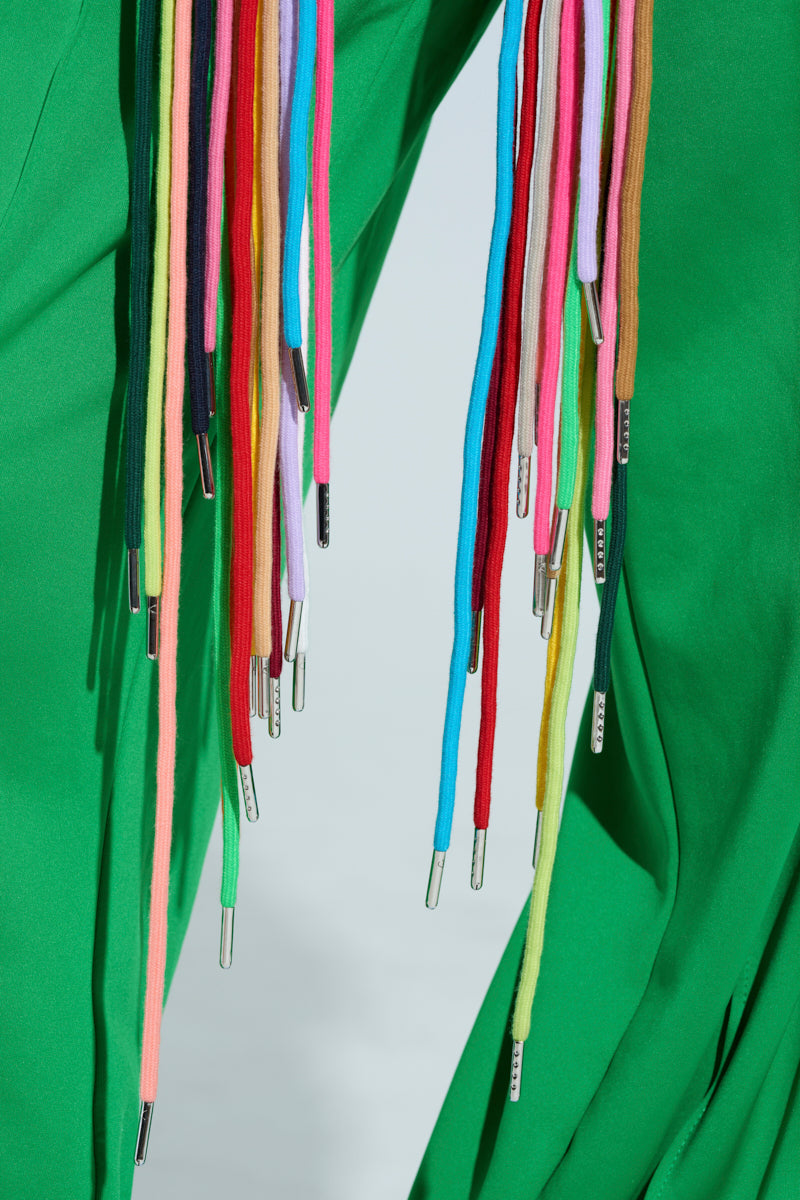 A close-up of someone wearing the STRINGED HOODIE in green, showcasing its colorful shoelaces, including yellow, pink, blue, and red with metallic aglets dangling against a simple backdrop.