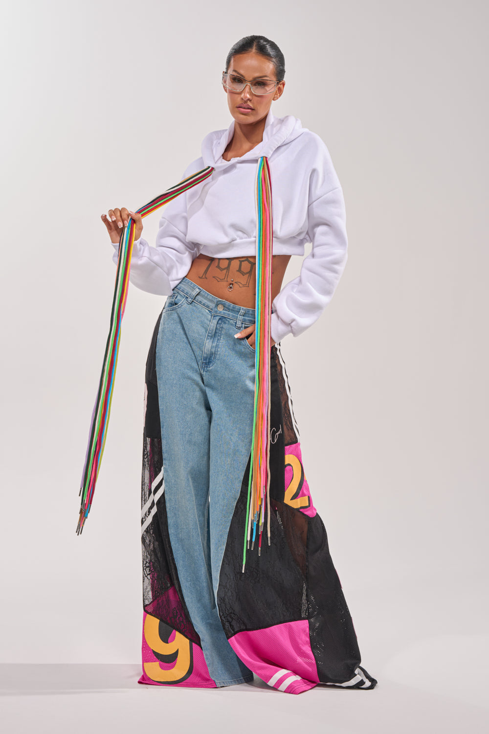 A woman in a STRINGED HOODIE, loose high-waisted jeans with colorful mesh panels and bold numbers, clear glasses, and long rainbow ribbons around her neck stands against a plain light background. 7