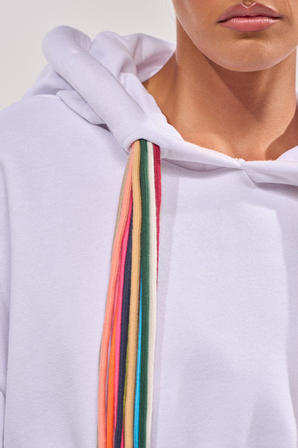 Close-up of a person wearing the STRINGED HOODIE, featuring multicolored drawstrings. The focus is on the vibrant strings set against the plain white fabric, emphasizing the hoodie’s streetwear style. 8