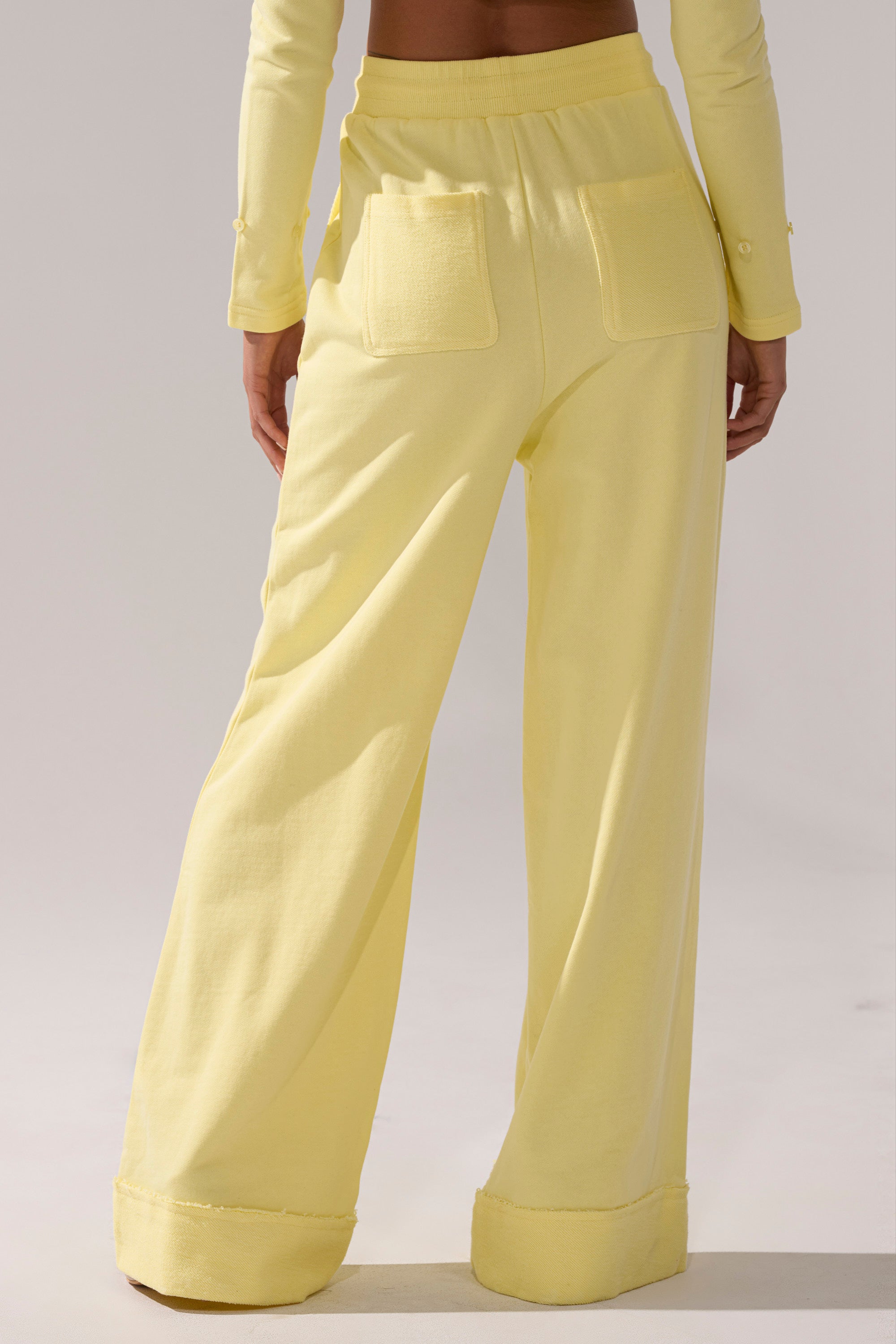 A person is photographed from behind wearing BUTTERFINGER WIDE LEG SWEATPANT in light yellow, featuring a high-rise fit and two back pockets, paired with a long-sleeve top against a plain light background.