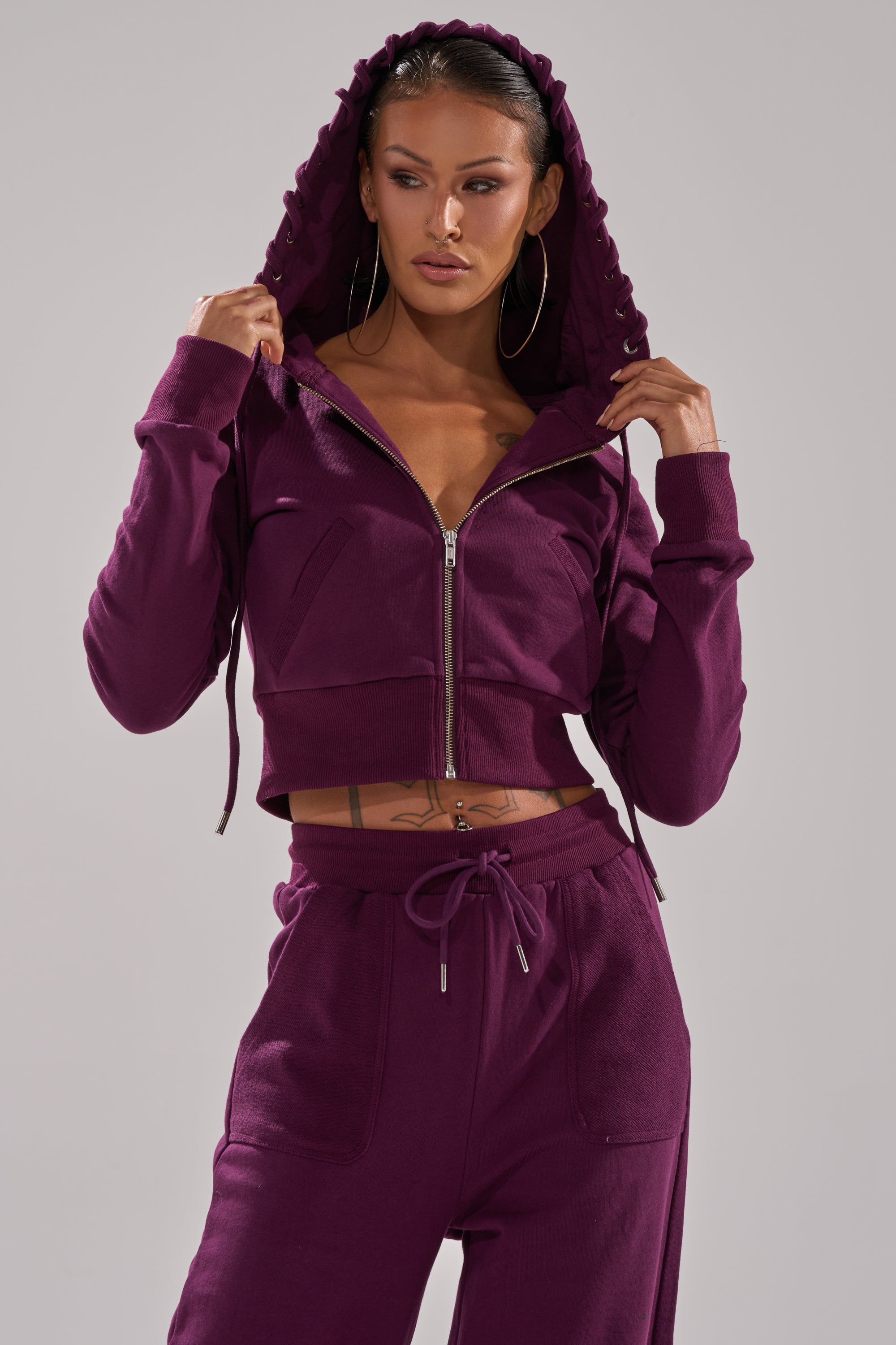 A woman stands confidently against a plain backdrop in the SHE'S GONE, YOU'RE FINE ZIP UP SWEATSHIRT—deep purple, unzipped with the hood up—paired with matching sweatpants, hoop earrings, and visible torso tattoos. 0