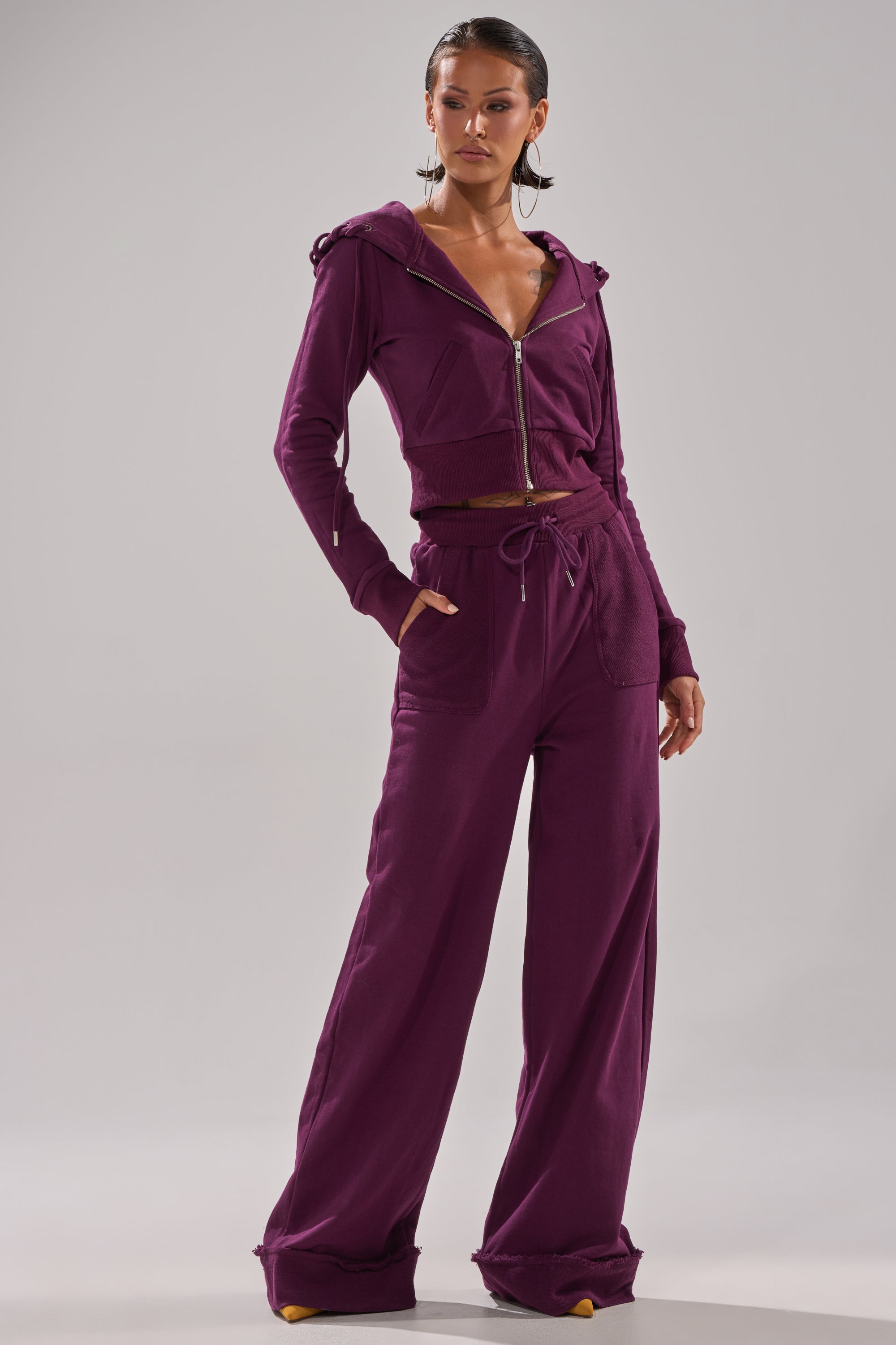 A woman poses in a chic deep purple SHE'S GONE, YOU'RE FINE ZIP UP SWEATSHIRT with wide-leg drawstring pants, standing against a plain light gray background. 9