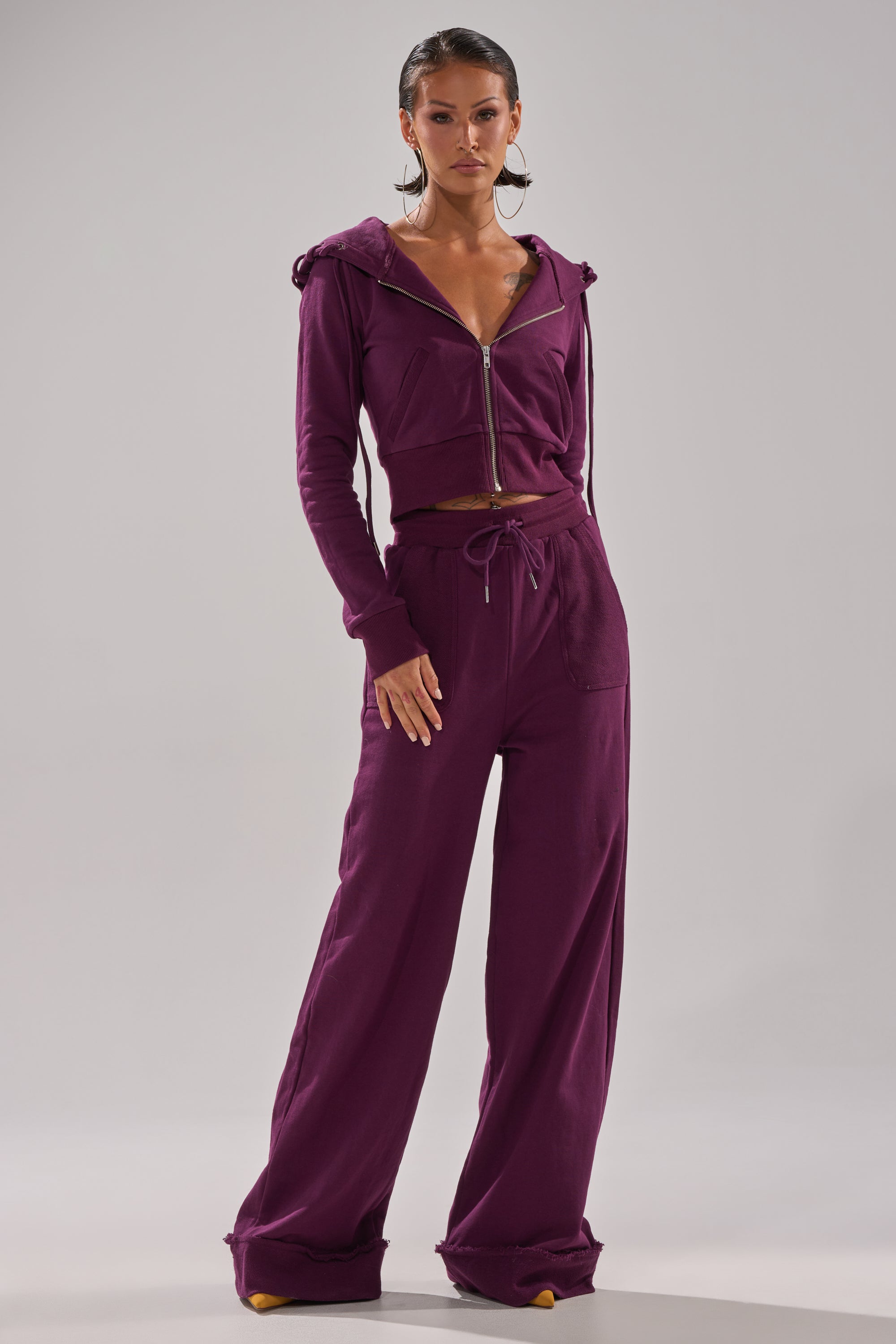 A woman stands confidently in a matching plum SHE'S GONE, YOU'RE FINE ZIP UP SWEATSHIRT and drawstring wide-leg pants, paired with yellow pointed-toe heels, against a simple light gray background. 10