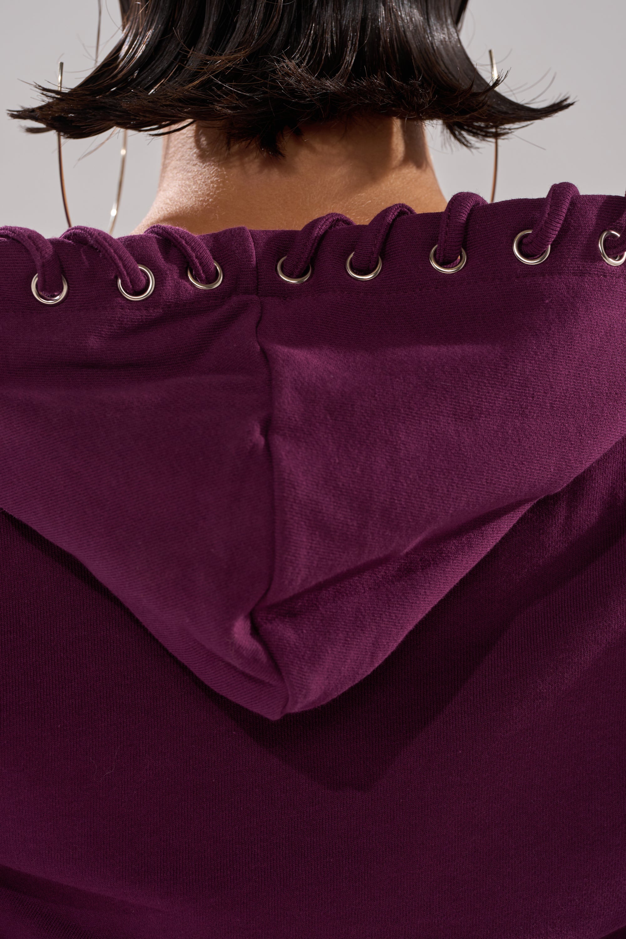 Close-up of the back of a purple SHE'S GONE, YOU'RE FINE ZIP UP SWEATSHIRT featuring metal eyelets and lacing along the hood. A hint of short dark hair and a large hoop earring are visible. 11