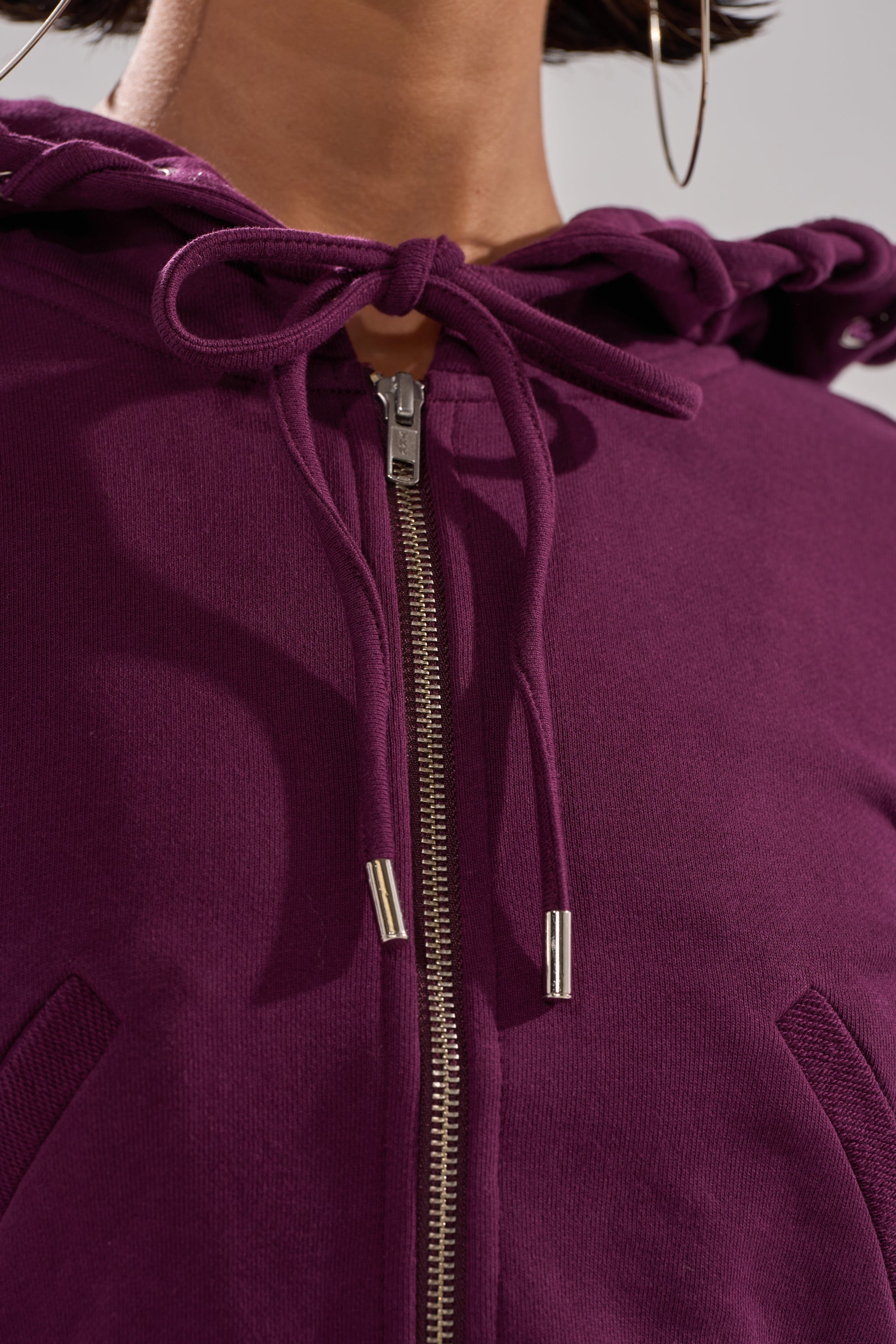 Close-up of a person wearing the SHE'S GONE, YOU'RE FINE ZIP UP SWEATSHIRT in purple. The zip-up style features a tied drawstring at the neck, partially closed zipper, and soft, textured fabric. 12