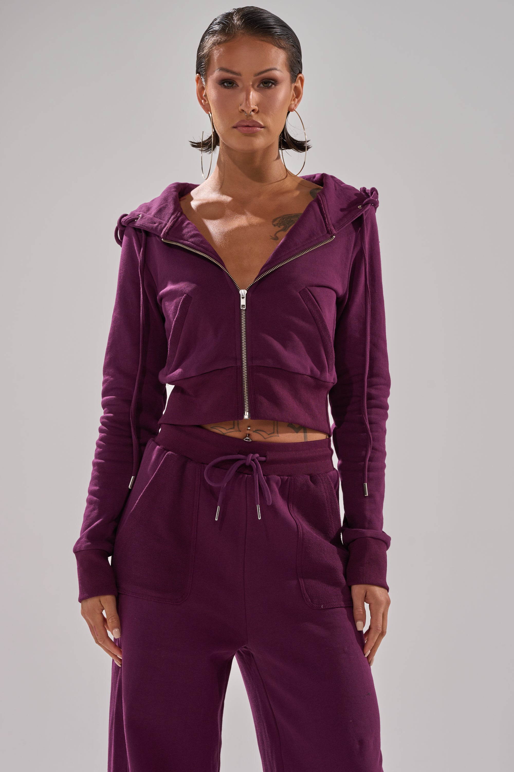 A woman with short, slicked-back hair and large hoop earrings stands facing forward against a plain background, wearing the SHE'S GONE, YOU'RE FINE ZIP UP SWEATSHIRT in deep purple with matching drawstring pants. 2