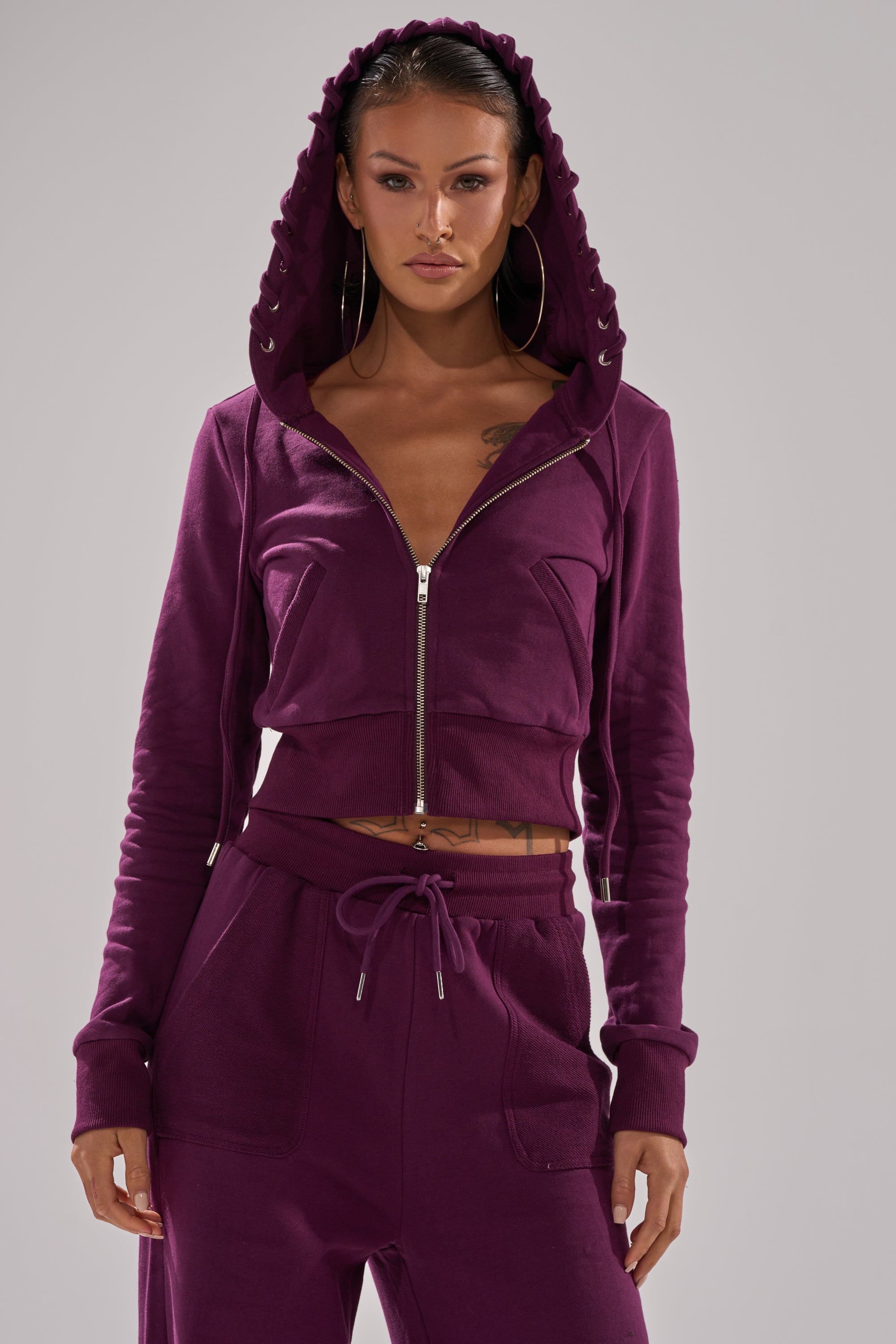 A woman stands facing forward against a plain light gray background, wearing the SHE'S GONE, YOU'RE FINE ZIP UP SWEATSHIRT—a purple cropped hoodie with the hood up and matching sweatpants, hands by her sides. 3
